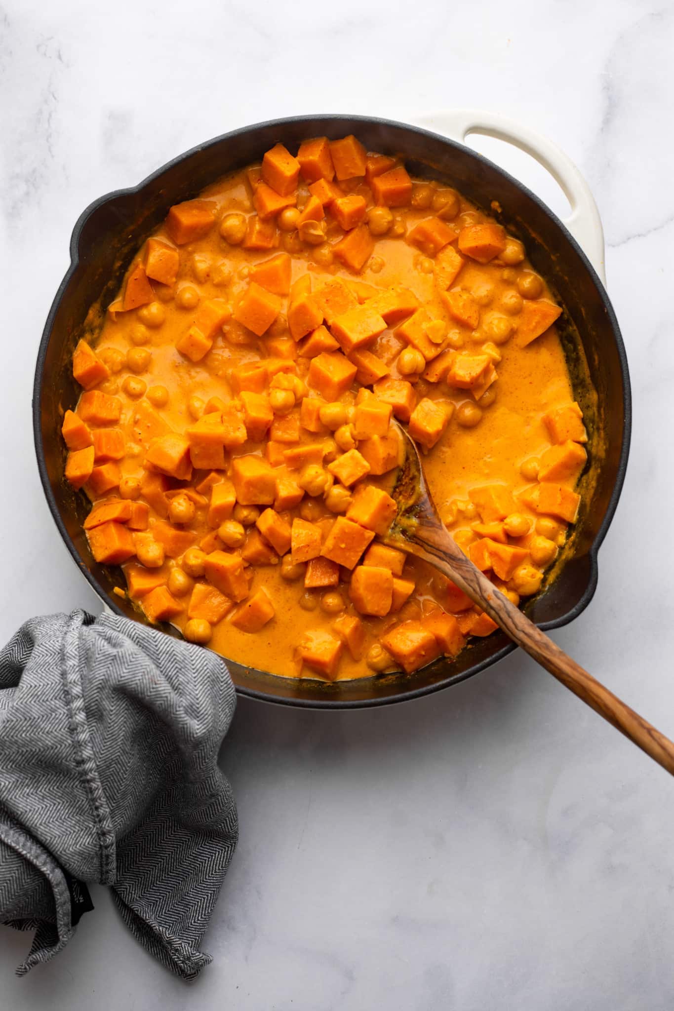 Stiring sweet potatoes in a red curry sauce with a wooden spoon in a black pan.