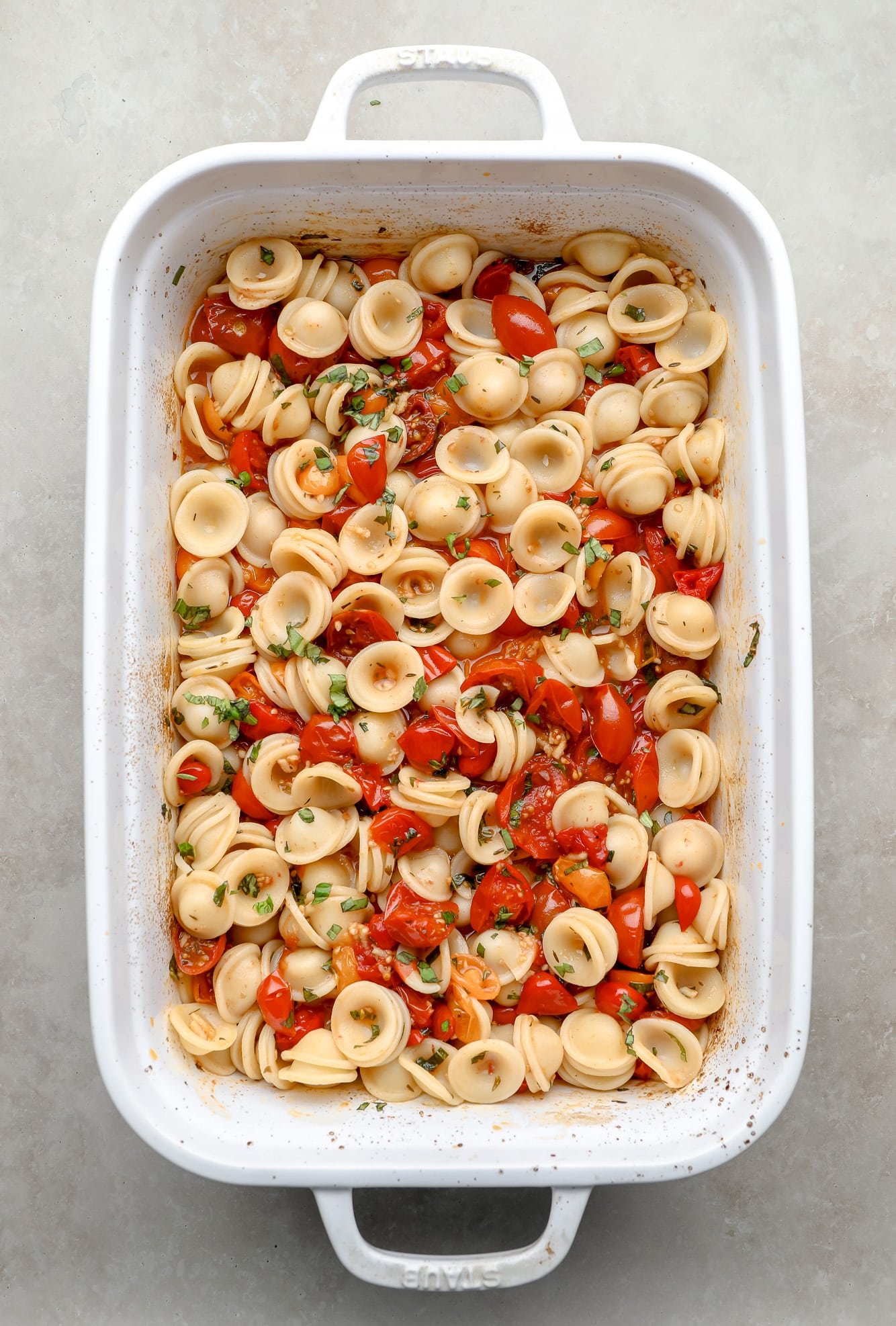 Pasta, roasted tomatoes and herbs joined in a white cooking dish.