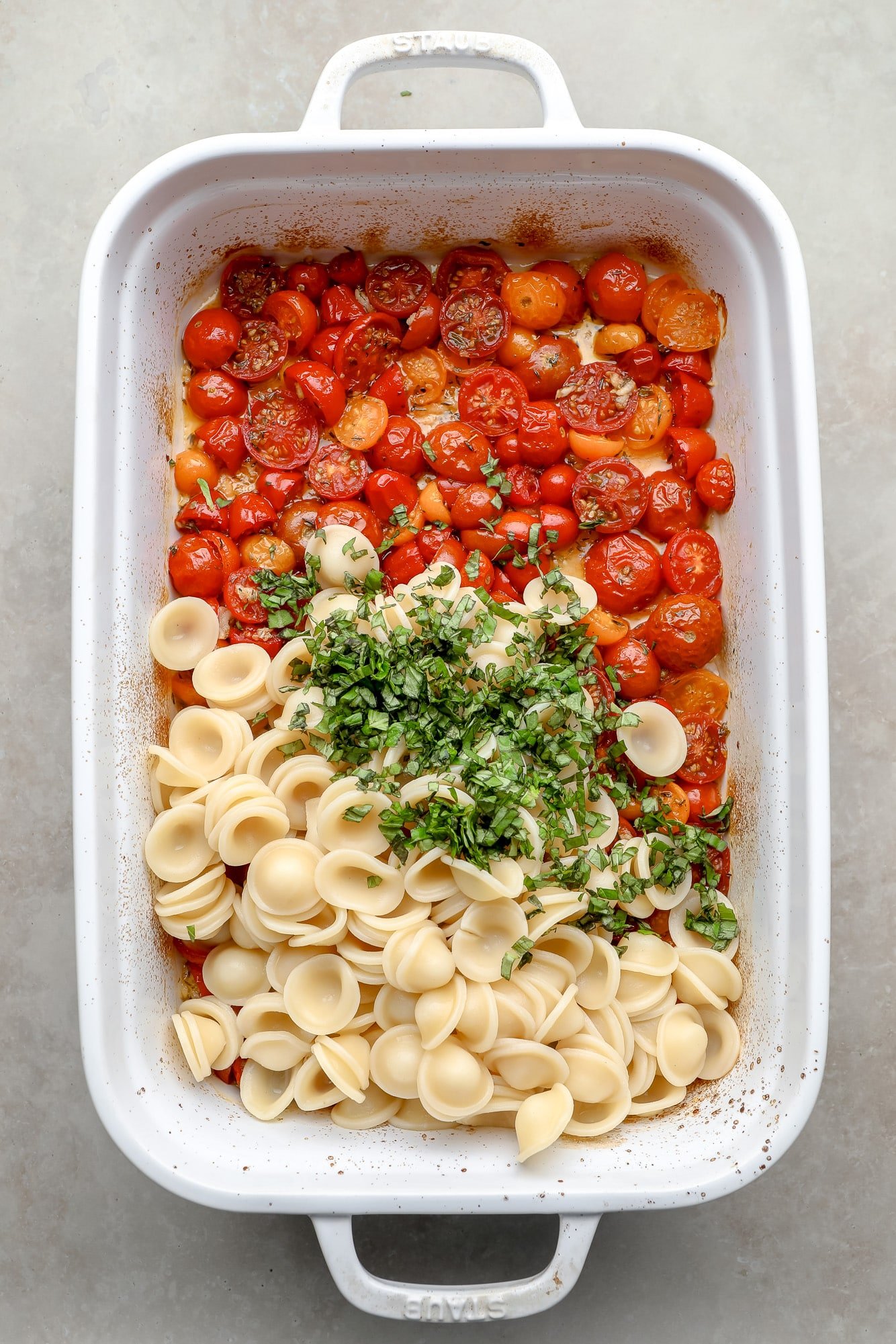 Cooked pasta and fresh herbs on top of the roasted tomatoes on a white cooking dish.