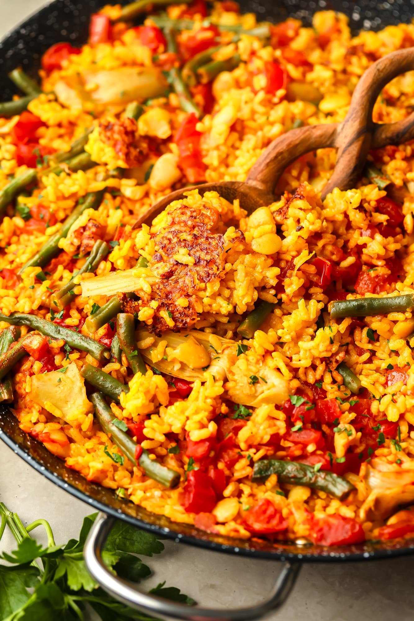 A wooden spoon lifting a spoon of vegan pan from a large pan of black paella.